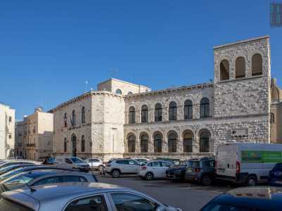 Oltre le auto si staglia il bianco prospetto della Corridoni, il cui aspetto imperioso rimanda subito alla sua epoca di costruzione. Fu infatti innalzata durante il fascismo, nel vivo dei lavori di risanamento di Bari vecchia